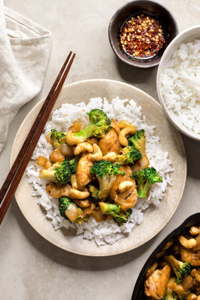 Gluten-free cashew chicken with broccoli on a plate with chopsticks, rice and chili flakes